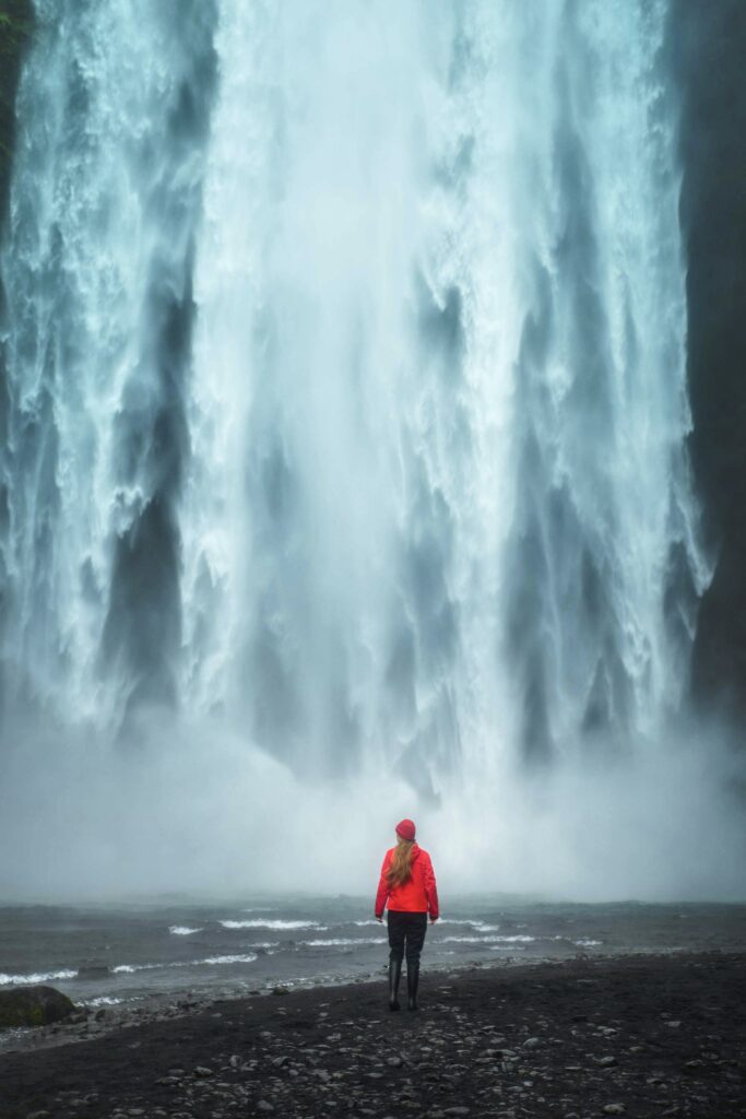 Woman in a red coat standing near the base of the massive Skogafoss waterfall in Iceland.