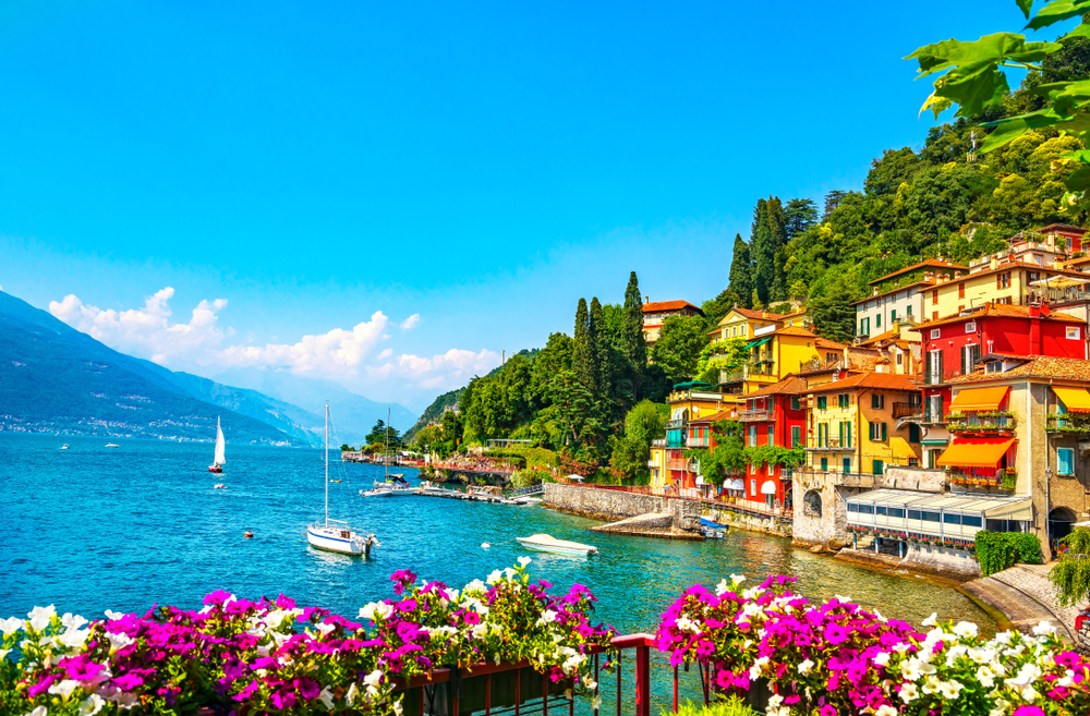 View of sailboats on Lake Como next to colorful buildings and flowers in the foreground.