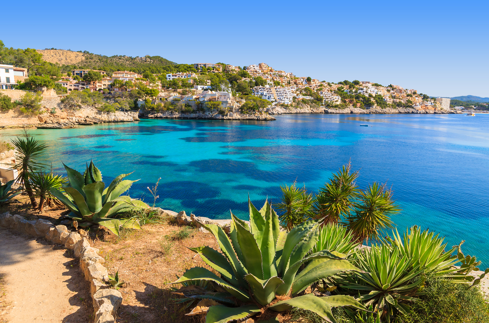 the view of the shore of the Island of Majorca. there are plants along the shore as well as apartments, condos, and villas. the water is clear blue and looks perfect for a swim. this is one of the best places to visit in Europe in April. 