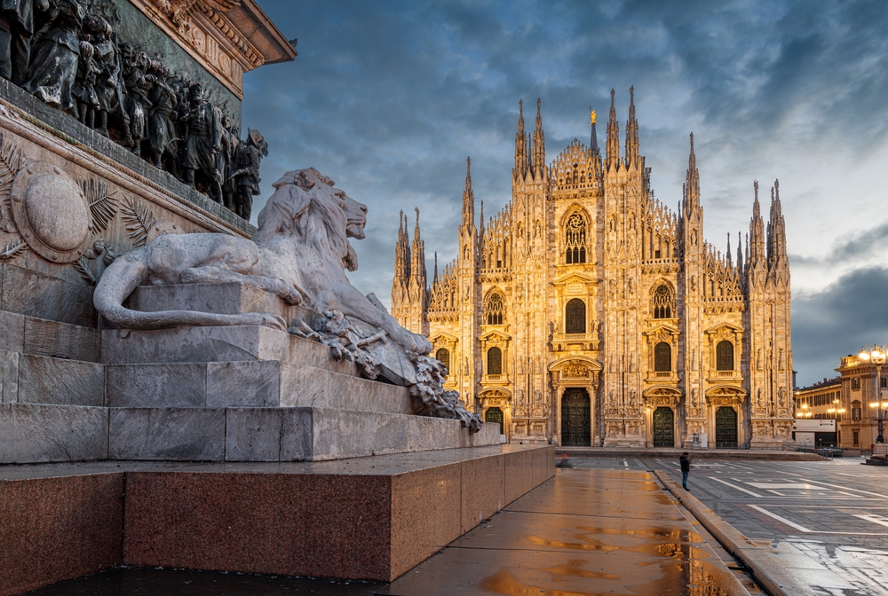 The Milan Cathedral lit up at dusk with a lion statue in the foreground.