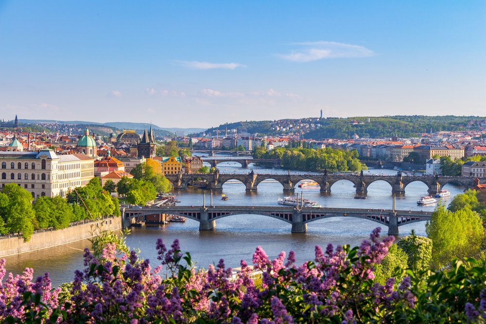 the view of Prague during the spring, there are flowers blooming and the photo shows at least 4 bridges and the city build along the mountains. Prague is one of the best places to visit in Europe in April 