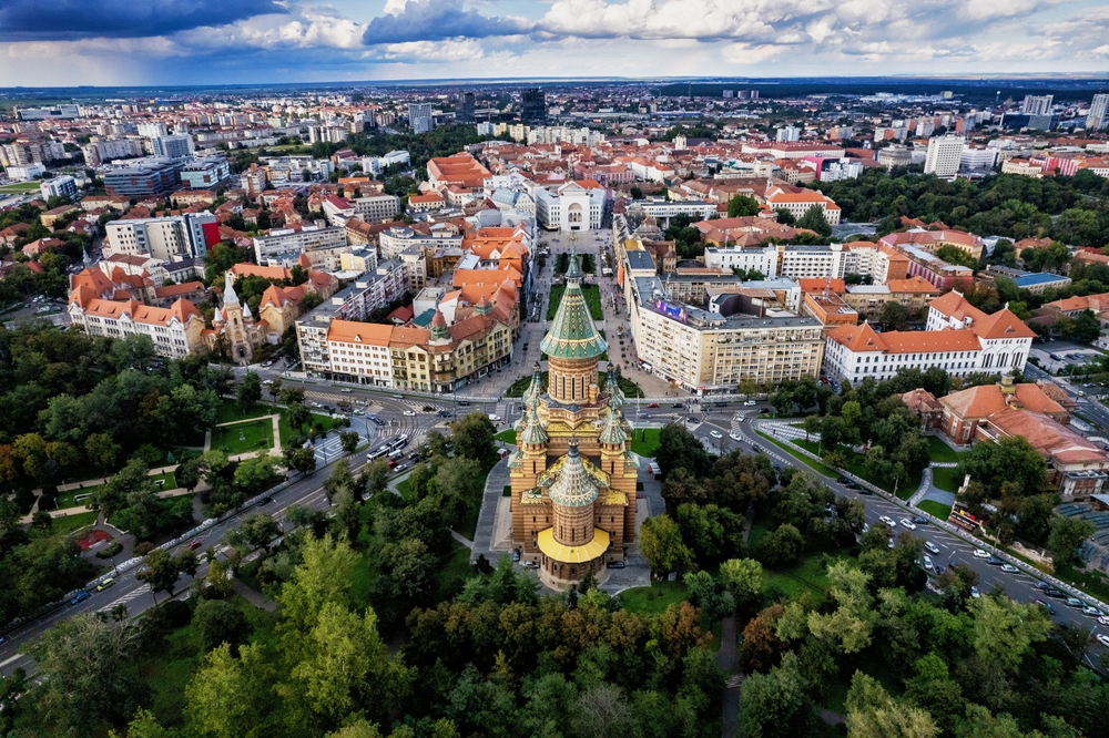 the beautiful town of Timișoara, Romania. there is a yellow and green church that over looks the city full of orange roof tops and a forest on the other side. 