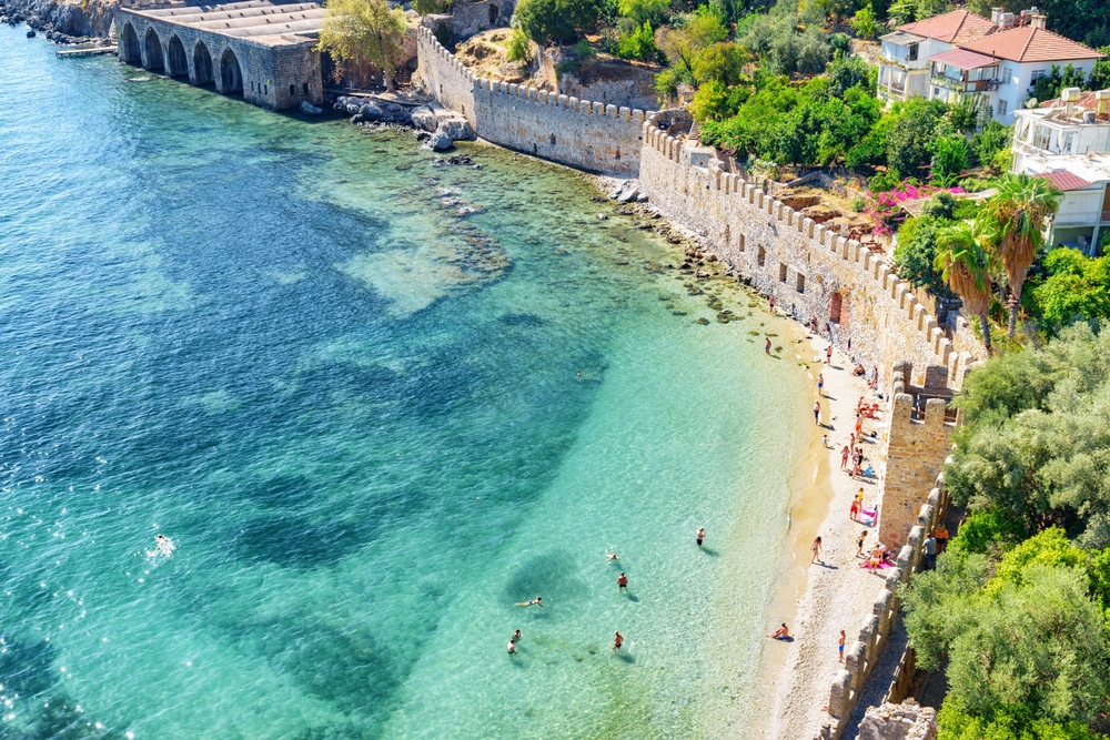 a photo of one of the towns on the Turkish Riviera. there is a castle wall protecting the town and pretty clear water with people swimming. 