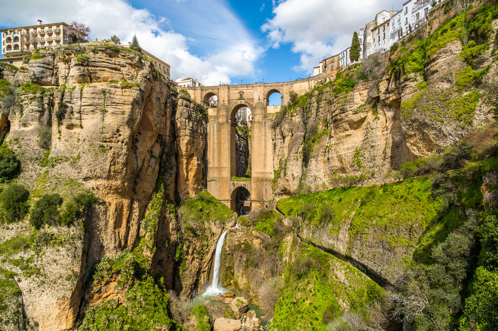 A unique bridge carved into a mountain with buildings around it and a waterfall below it. 