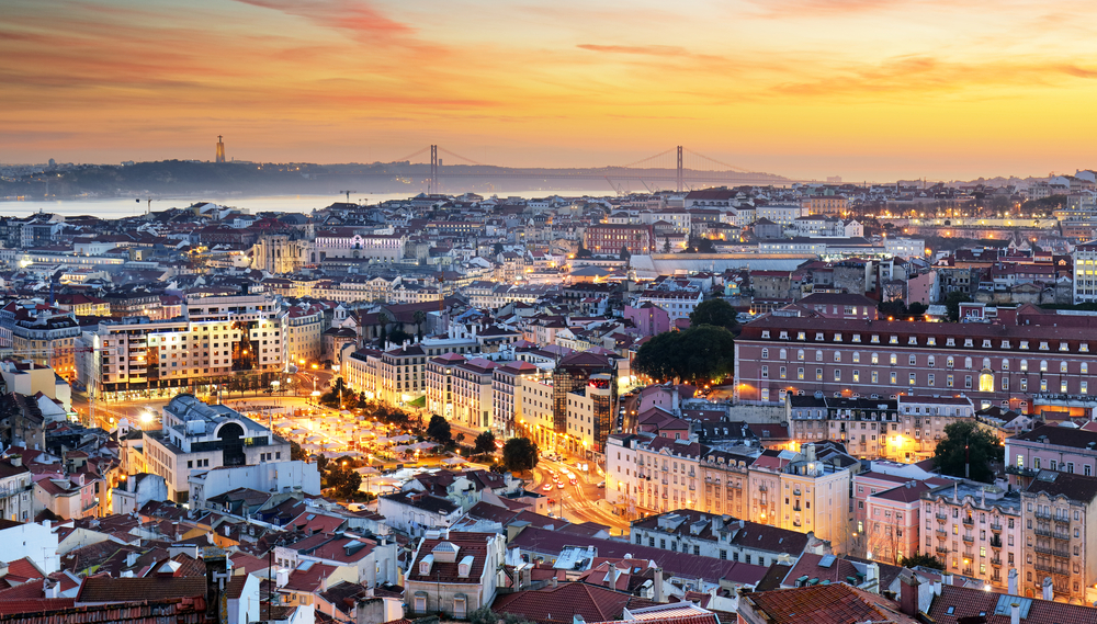 a few of lisbon portugal from the sky at sunset. you can see the epic bridges and the street lights glowing orange and bright, just like the sky at sunset 