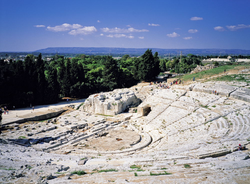 the view of the Neapolis Archaeological Park in Sicily during the winter time. this is an ancient theater from roman times. you can see a forest and mountains in the back around and it looks clear and blue in the sky 