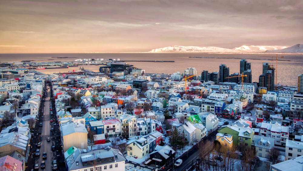 a beautiful photo of Reykjavik Iceland during February at sunset. there are snowy mountains in the back around and the town has cute family homes and big condos. 