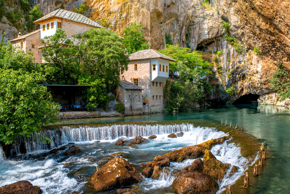 a curved waterfall cascading in front of river front buildings, trees, and rock cliffs in Bosnia and Herzegovina 