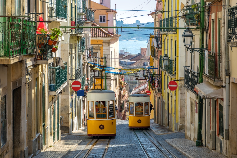 two cable cars in the streets of portugal, stop signs are on both sides of the photo and there are wires above the cable cars, water in the distance 