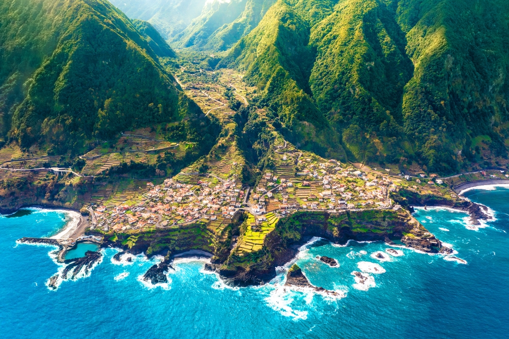 aerial photo of madeira with waves crashing on the coast and mountains in the background, one of the best palces to visit in europe in march 