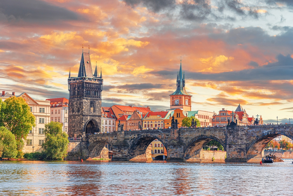 an arched bridge  over a river in front of historic buildings and a clock tower, there are people standing on the bridge