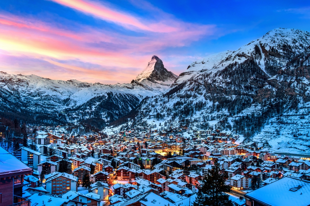 a snowy mountain village between mountain peaks, lights are shining in the buildings at sunset 