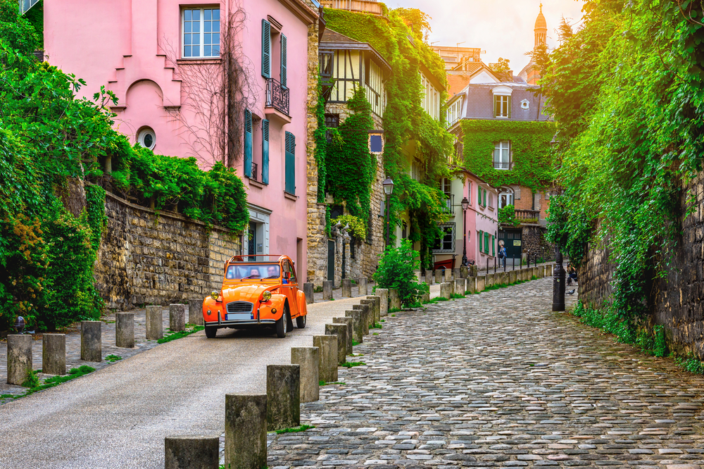 a car drives on historic road next to row of houses, cobblestone are on the right 