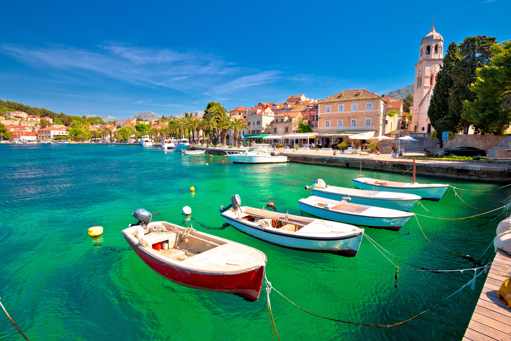 Typical Croatain flat bottom boats floating on the water in one of the towns on the coast. There are beautiful palm trees, churches and mountains in the back round. 