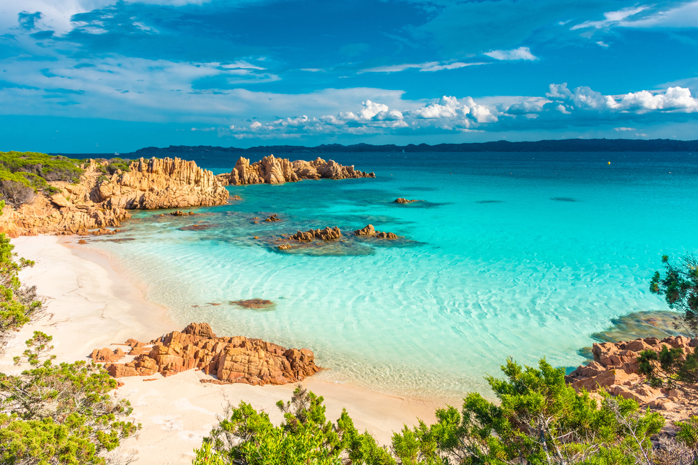 a beach on the Sardinia island in italy . the water is a beautiful clear blue tone and there are sea mountains that are a pretty orange. 
