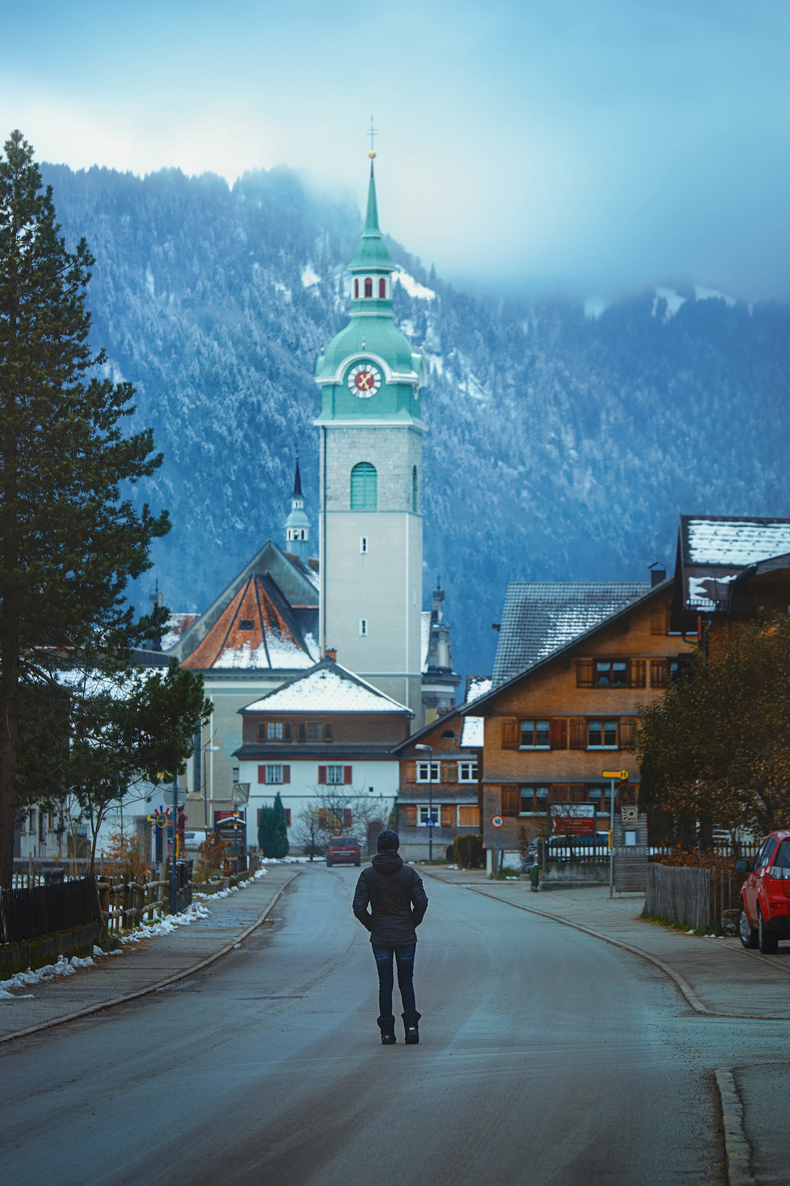 Girl walking along a European street there are snow covered streets There is a forest in the background. 