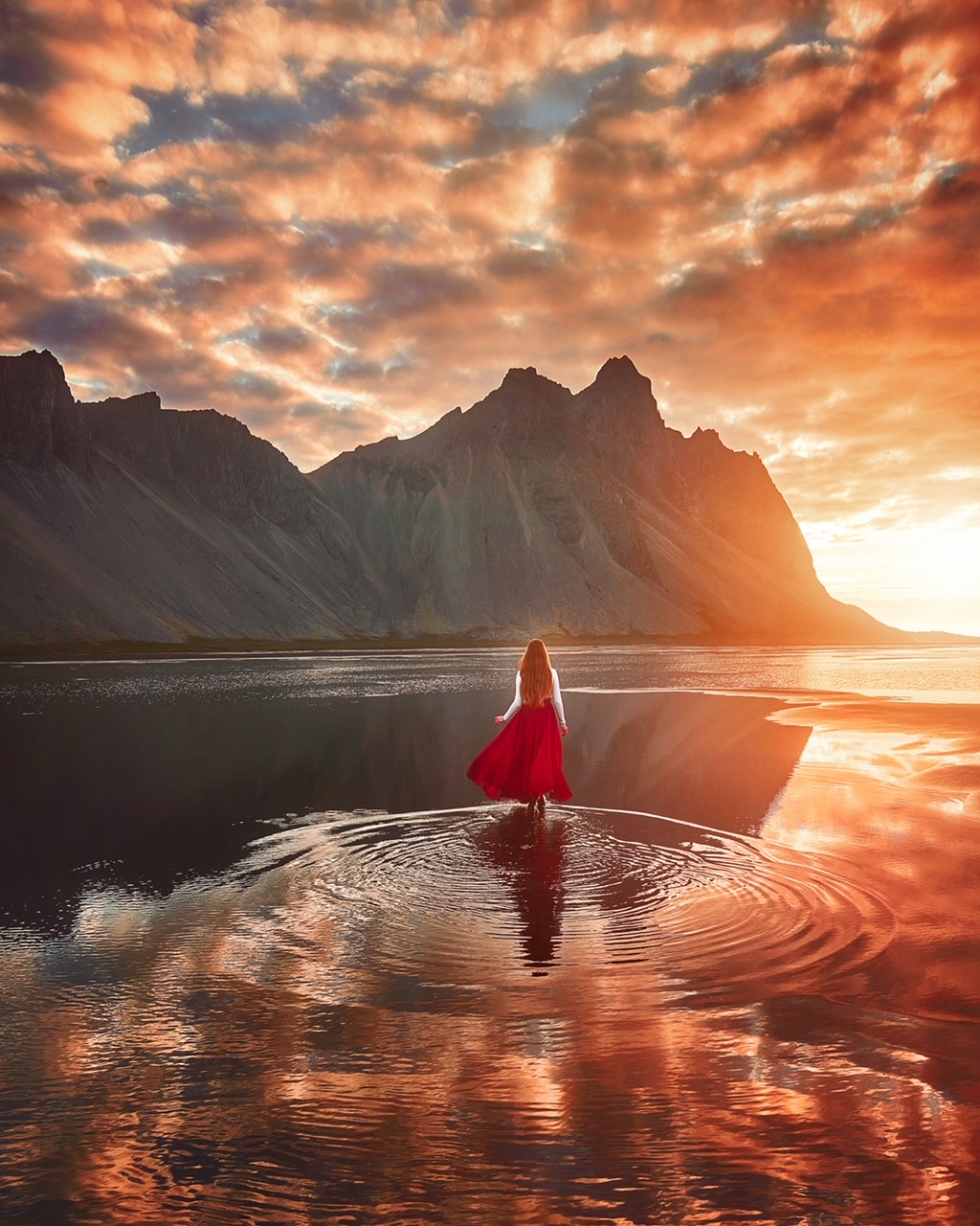 Girl on a beach in Icleand with the sun setting. She has a red skirt on and their are cliffs in the distance. 