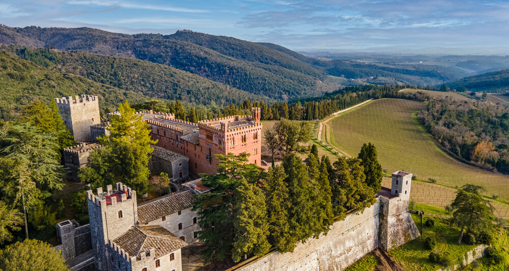 Tuscan castle with brown stone perched on a cliff with wine fields below and green mountains in the background