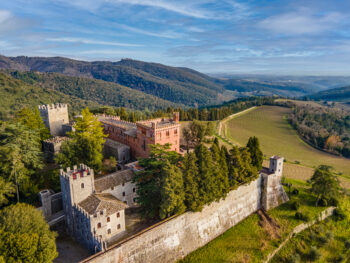 Tuscan castle with brown stone perched on a cliff with wine fields below and green mountains in the background