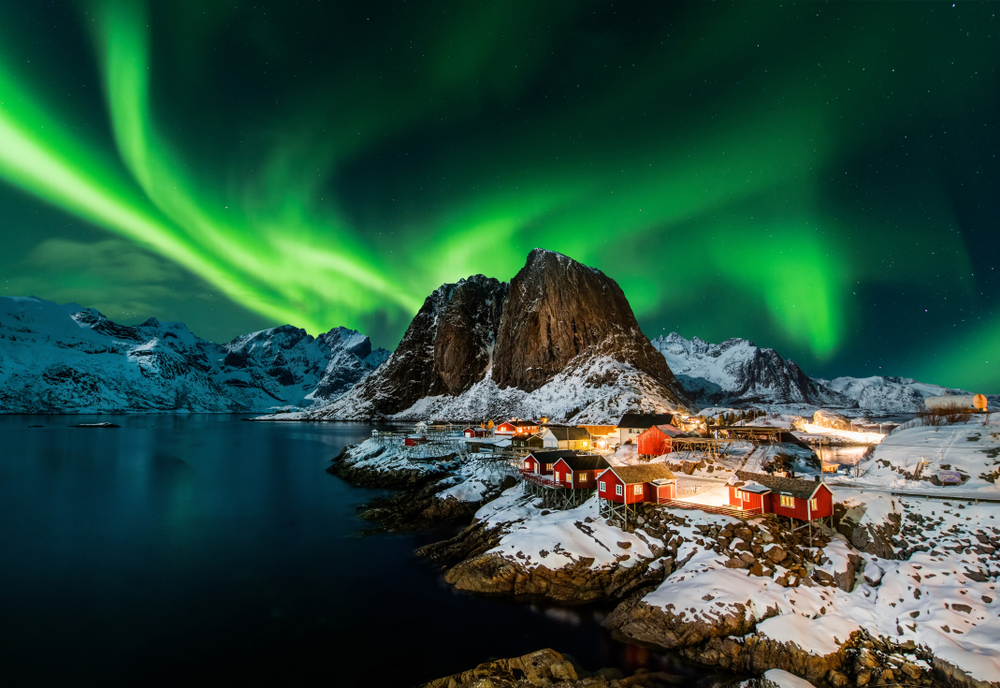 A small fishing village with red buildings, some snow on the ground, a mountain in the distance, and the Northern Lights in the sky, at night