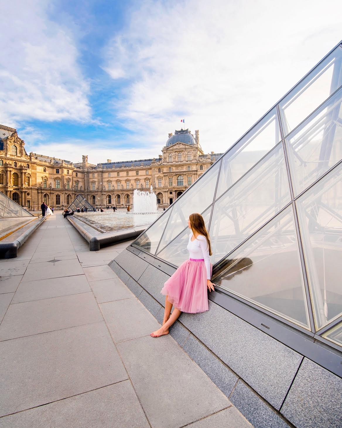 The Louve can't be missed on your Paris in 2 days Itineray a girl in pink skirt is sitting outside the glass pyramids outside the museum