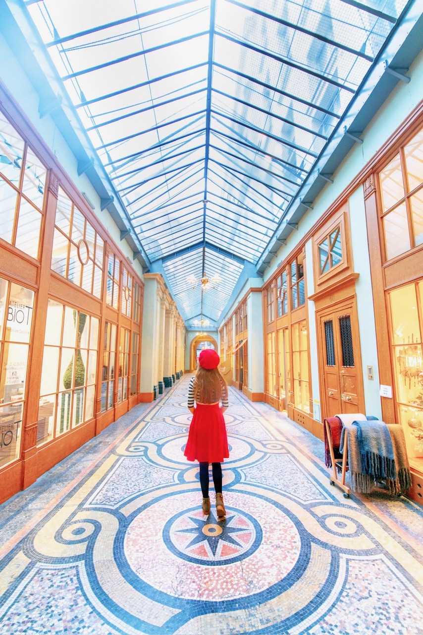 A girl in Red skirt and red beret is standing on the mosaic floor of one of the covered passageways inn Paris filled with light and glass