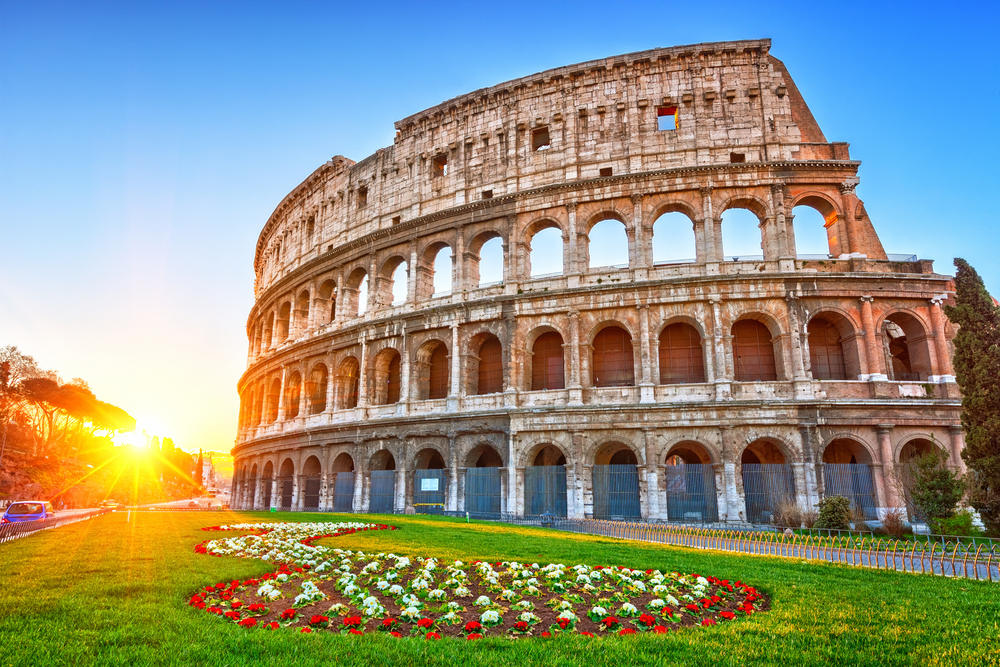 the rome colosseum during spring with the sun setting in the corner, there is a big patch of spring flowers and nobody around, rome is one of the best places to visit in Europe in April 