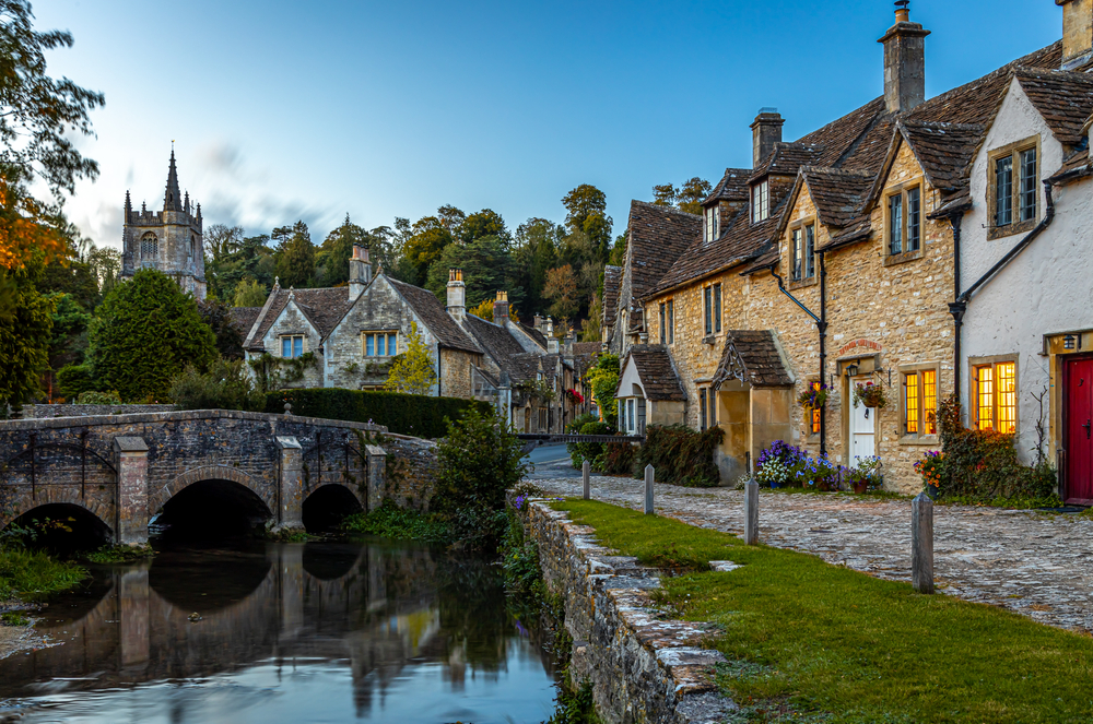 View of Castle Combe one of the places in southern England. There are old houses and a bridge over a river. You can see a church spire in the background.  