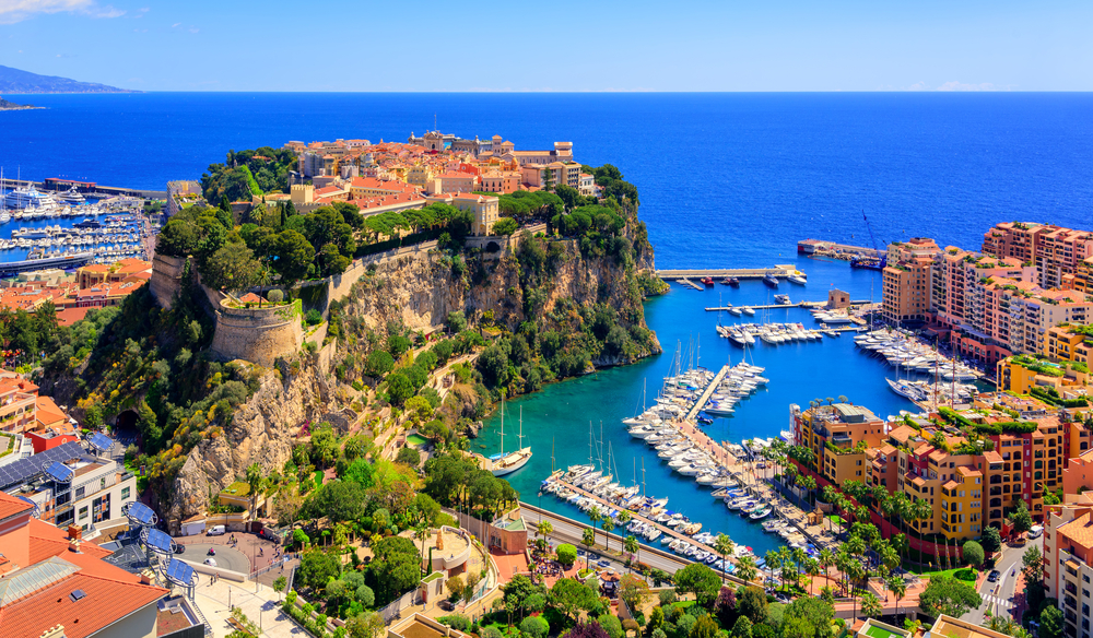 A view of a bay on the side of the ocean with rocky cliffs, boats docked at a marina, and tall terracotta colored buildings. 