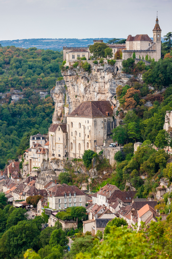 the cliff town of Rocamadour. You can see buildings on the cliff and at the bottom. The town is surrounded by trees. 
