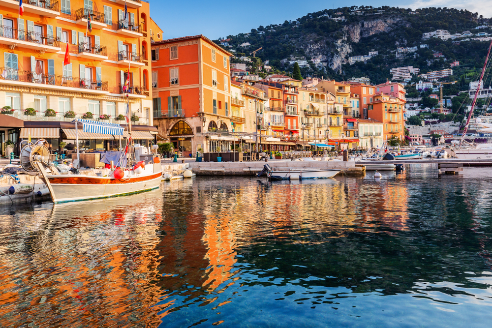 Seaside town on the French Riviera showing boats in front of colorful houses with a mountain on the background. 