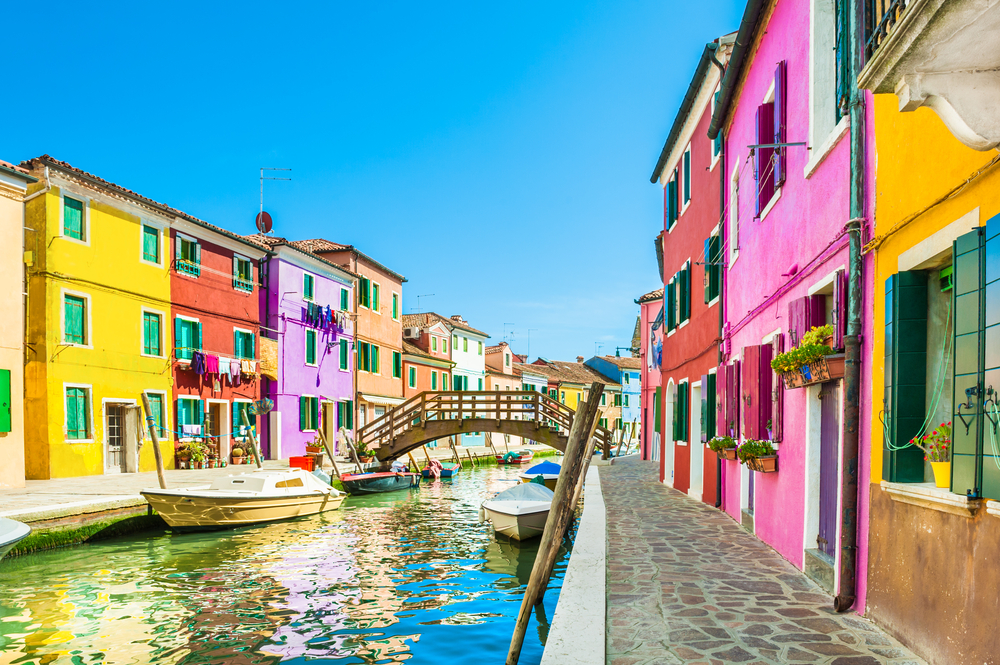 Bright, colorful buildings along a canal with a wooden bridge and boats in Burano, Italy.