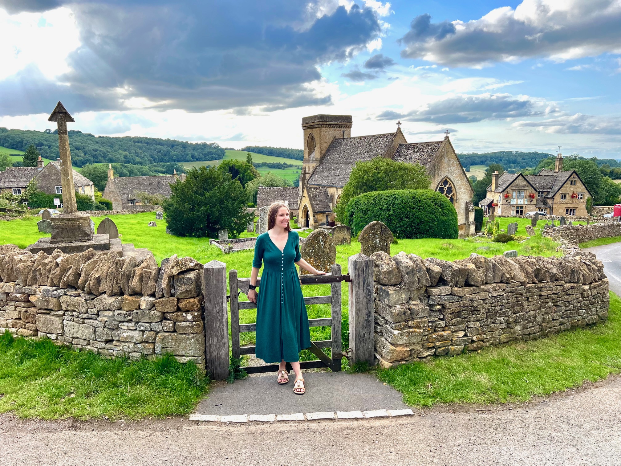 Woman in a long green dress standing by the gate of a graveyard by a church in the Cotswolds.