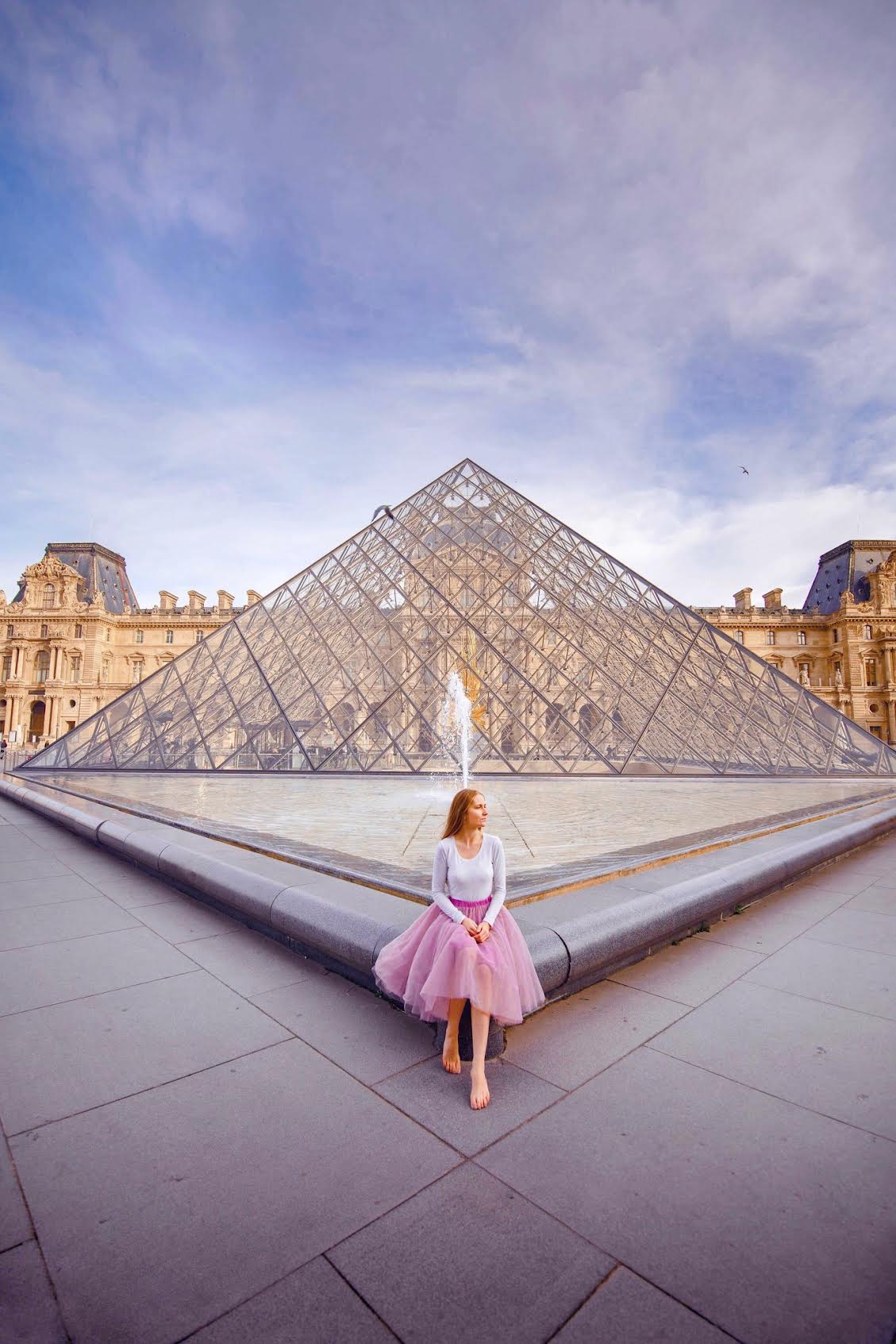 Woman in a pink tulle skirt sitting on the edge of the fountain next to the glass pyramid of the Louvre Museum in Paris.