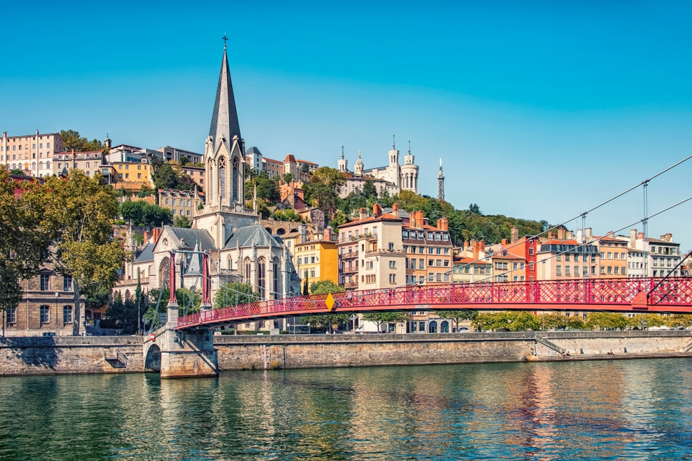 A red, metal bridge going across a river near a beautiful church in the city of Lyon.