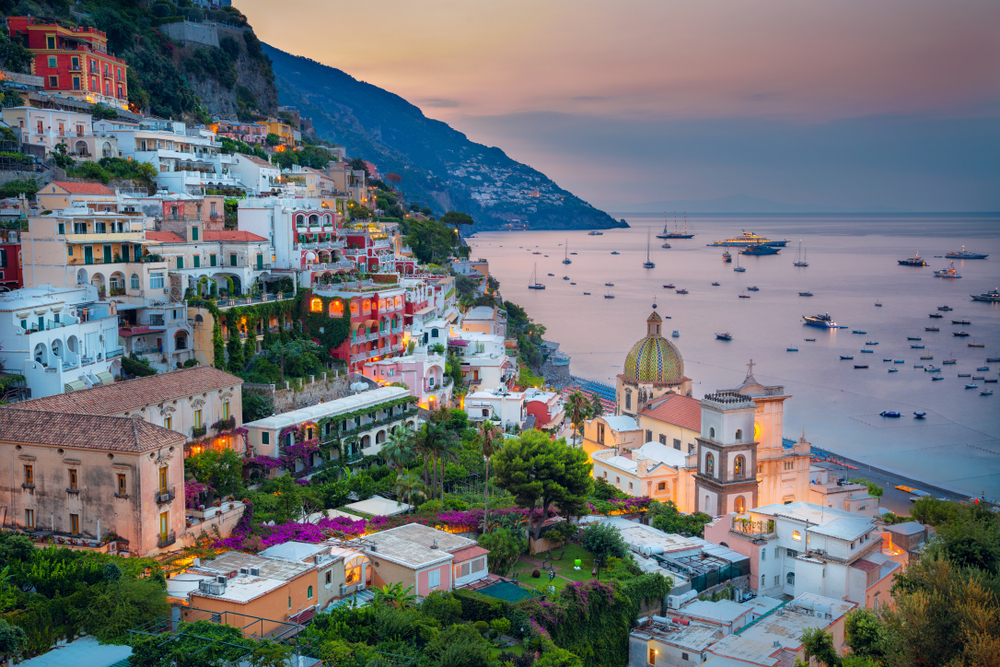 View looking down at Positano during sunset with the buildings lit up and many boats on the water.