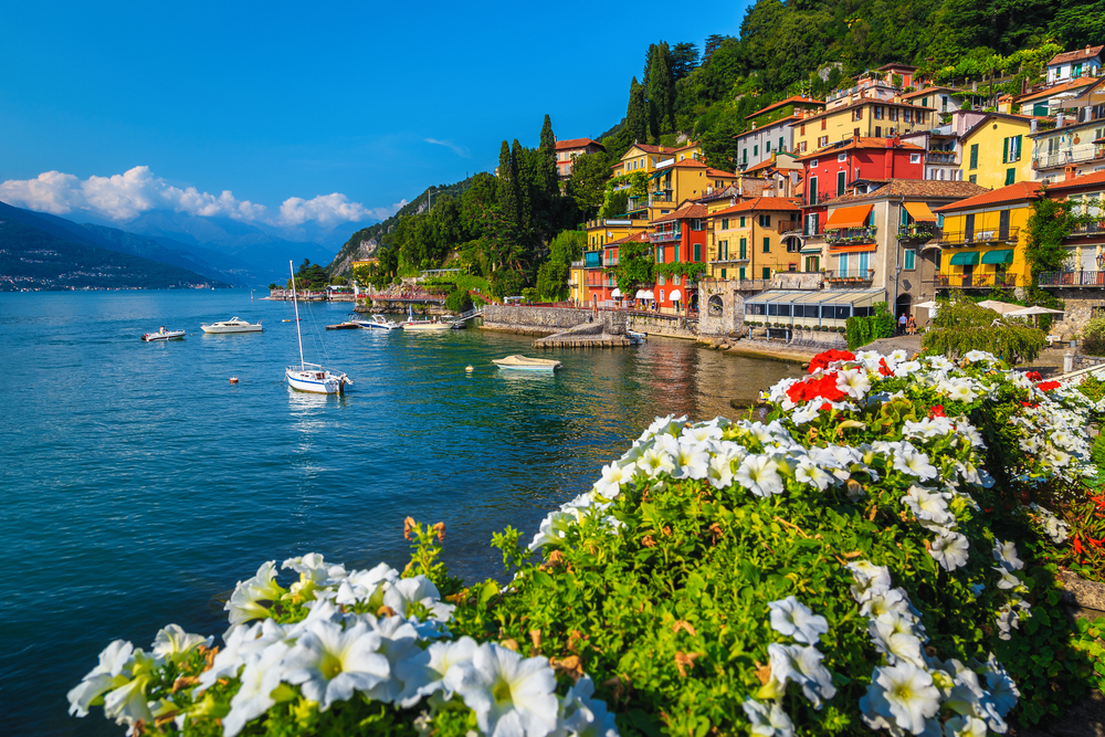 Colorful buildings along the water with boats with flowers in the foreground in Varenna, one of the best small towns in Italy.