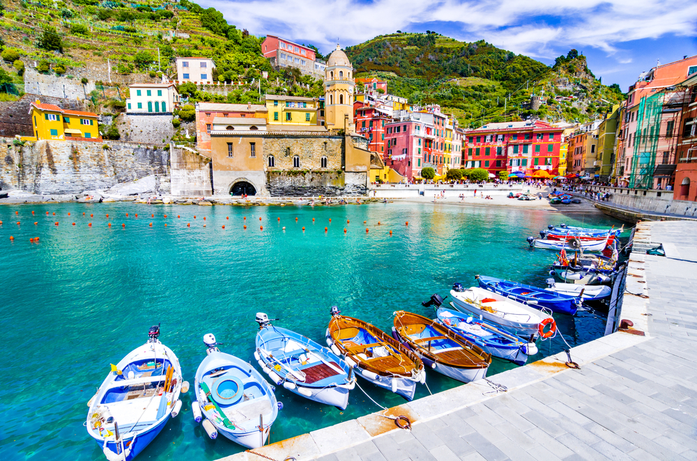 Boats docked in a bright blue harbor near a beach and colorful buildings in Vernazza.