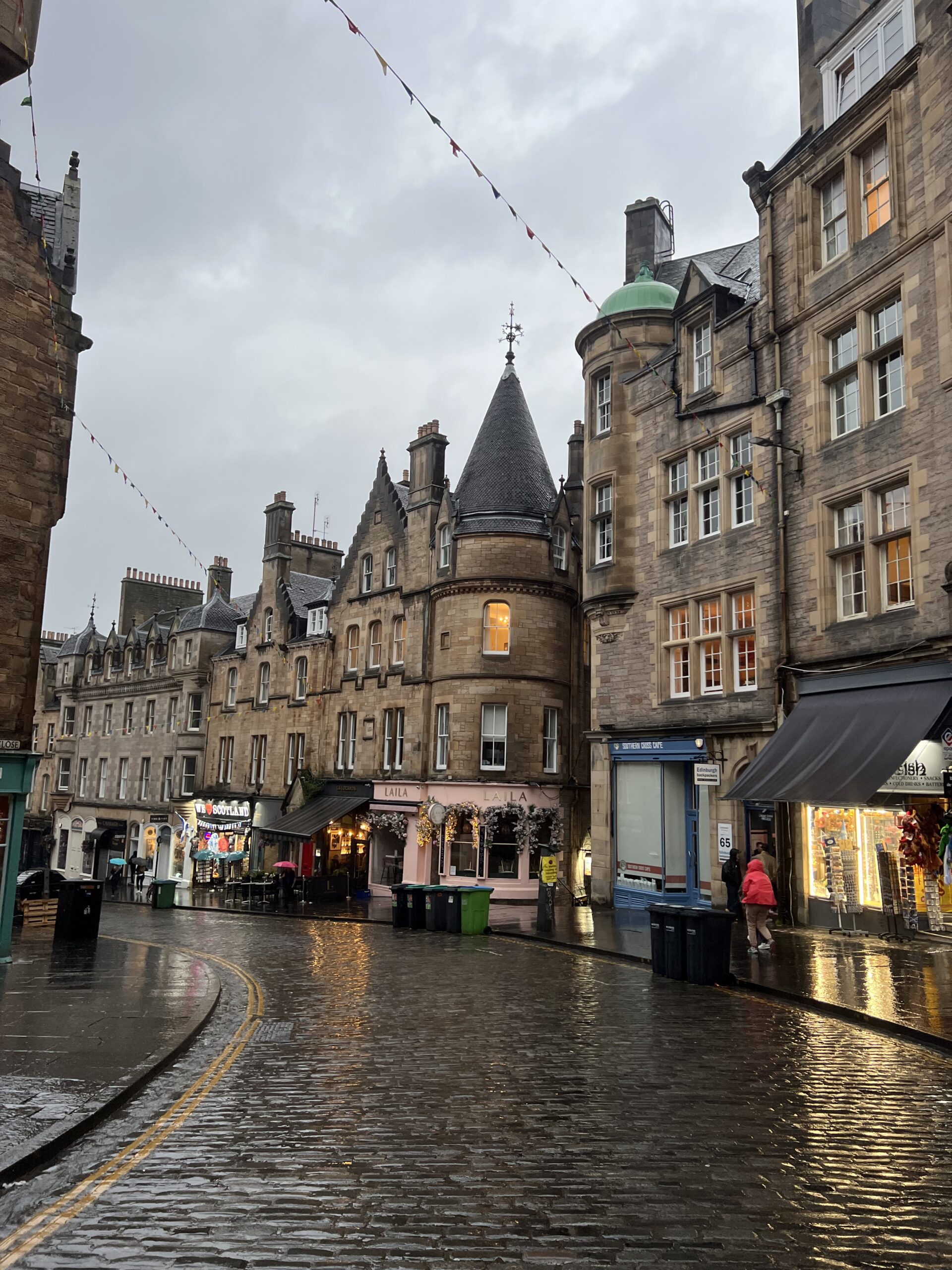 a photo of a wet street in Edinburgh Scotland where people are walking along the street in front of shops and restaurants 