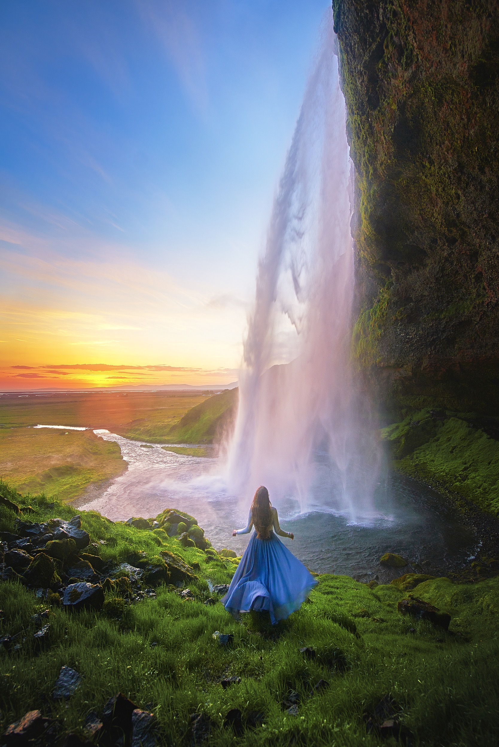 a woman is standing in a long dress in front of a pool of water and a tall waterfall at sunset , one of the best places to visit in Europe in July