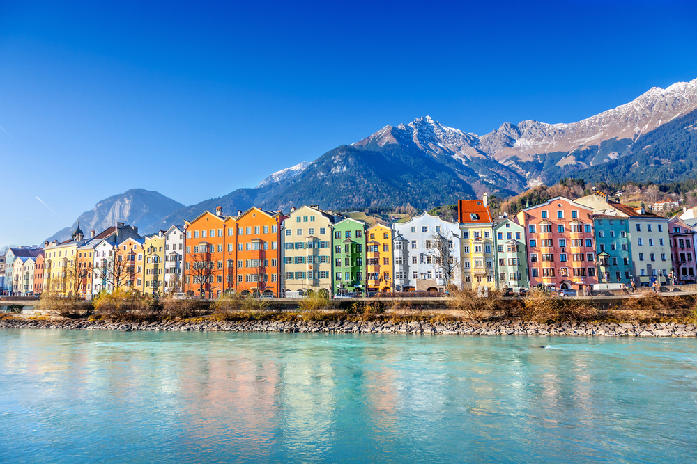 a row of colorful houses sit on the waters edge with mountains behind them, photo was take on a clear day with no clouds in the sky 