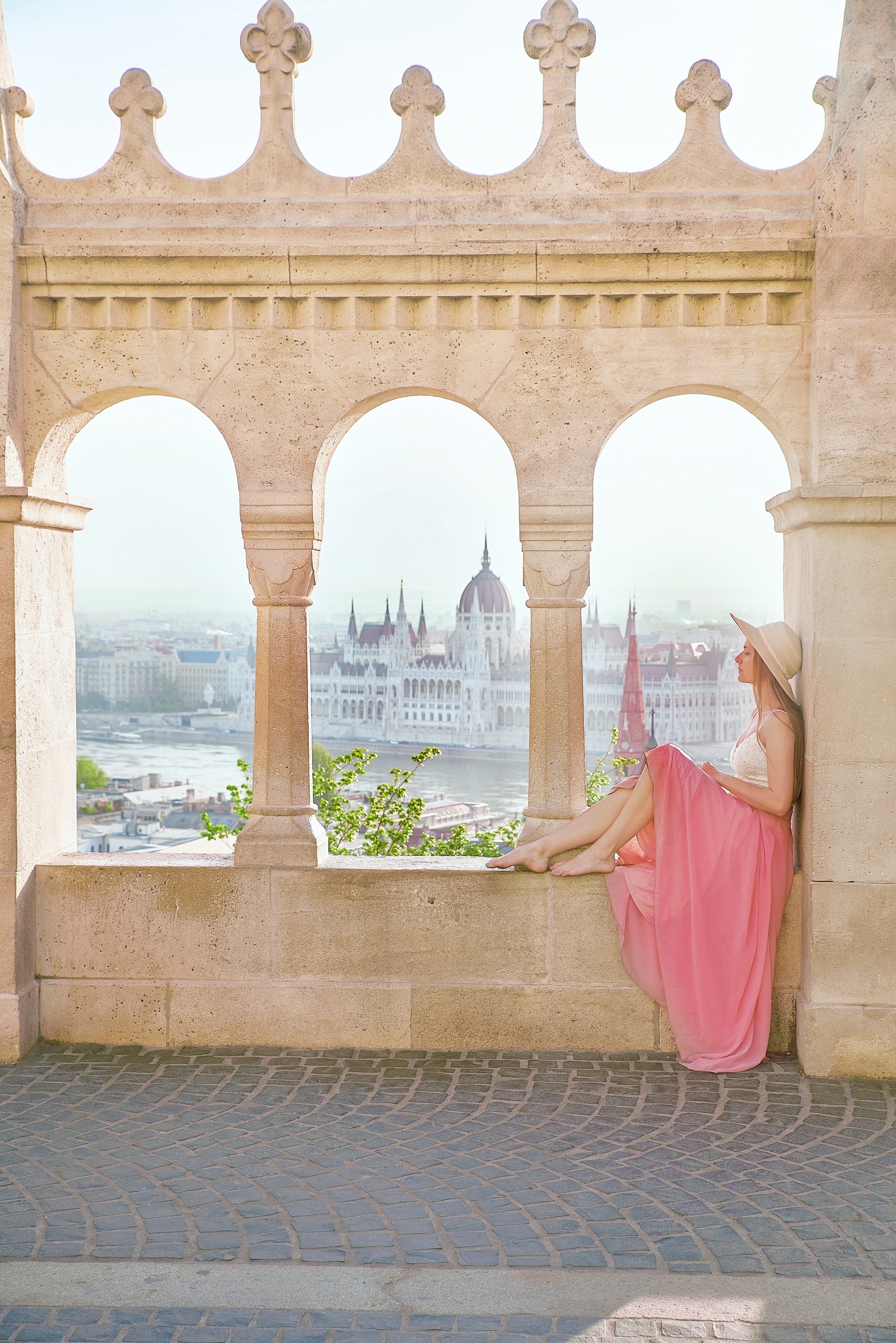 a woman sits in an archway with her feet propped up overlooking the river and city below, she is wearing a long dress and a hat