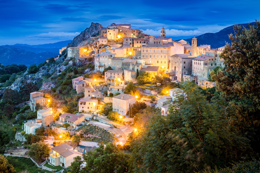 stone buildings sprawl over a hill in corsica at dusk, lights are on outside the buildings in the streets