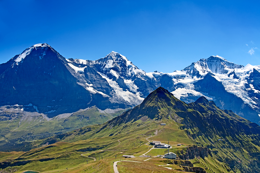 mountains with snow on them are in the background, in the middle of the photos there are grassy mountains and hills and in the foreground there is a grass field with a walking trail in the middle of it 