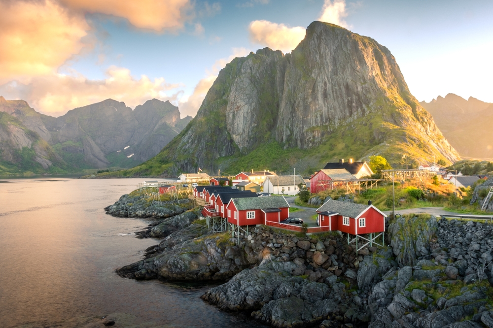 one of the best places to visit in Europe in July, red houses sit on waters edge in lofted islands Norway with rock formations in the background 