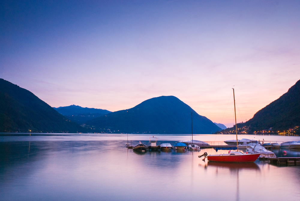 sunset mountain backdrop and glassy waters on the beautiful Lake Lugano Italy. There is boats on the water. 