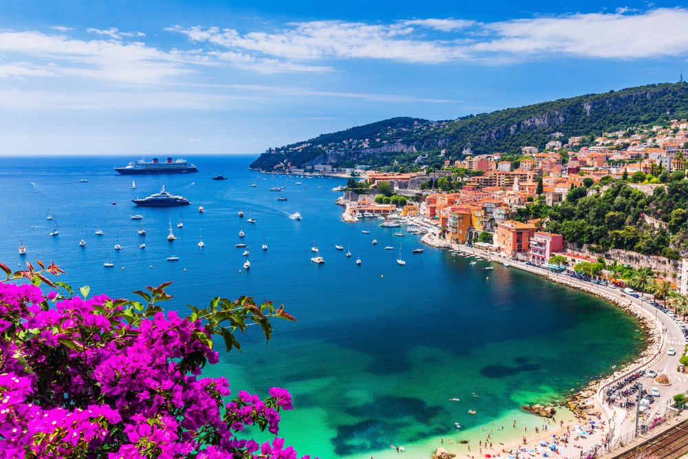 photo looking down on sandy beach on the Mediterranean Sea, bright flowers are on the left and on the right there are buildings an mountains, in the water there are many boats and cruise ships