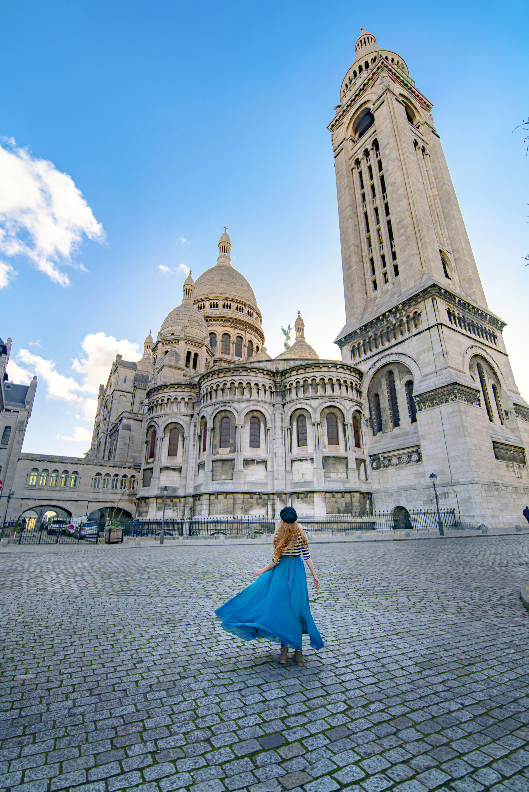 Girl in a long blue skirt in front of the Sacre-Coeur. She stands on a cobbled road in front of it. 