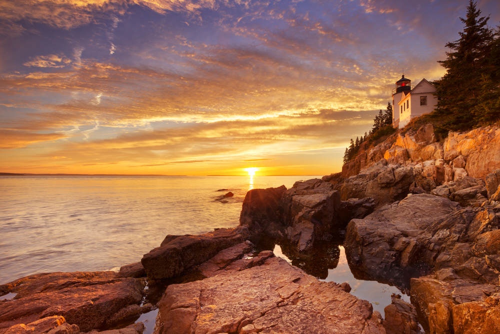 Vivid sunset over the ocean and a lighthouse on a stony shoreline in Acadia National Park on an East Coast road trip.