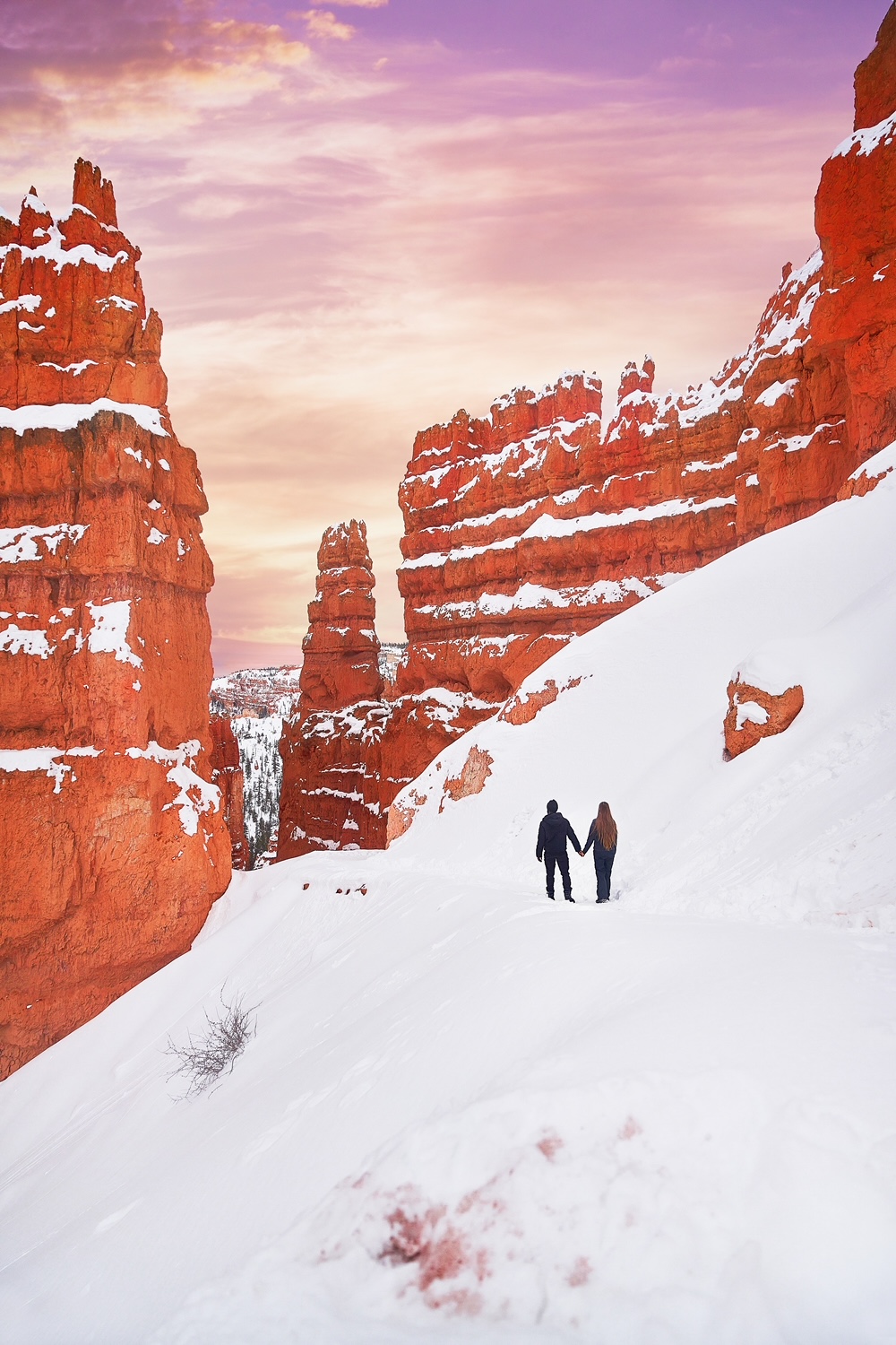 Couple holding hands as they hike through snow among hoodoos at sunset on a Utah road trip.